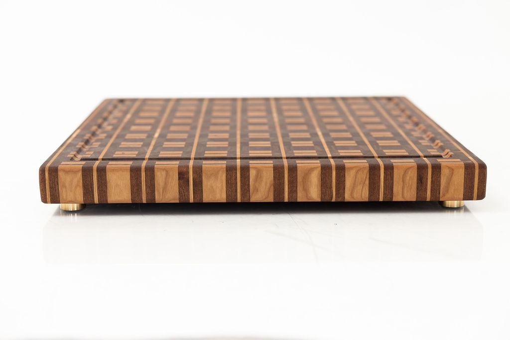 Plaid pattern end grain cutting board with brass feet, featuring complex geometric design in sapele, walnut, and cherry wood with juice groove and rounded corners